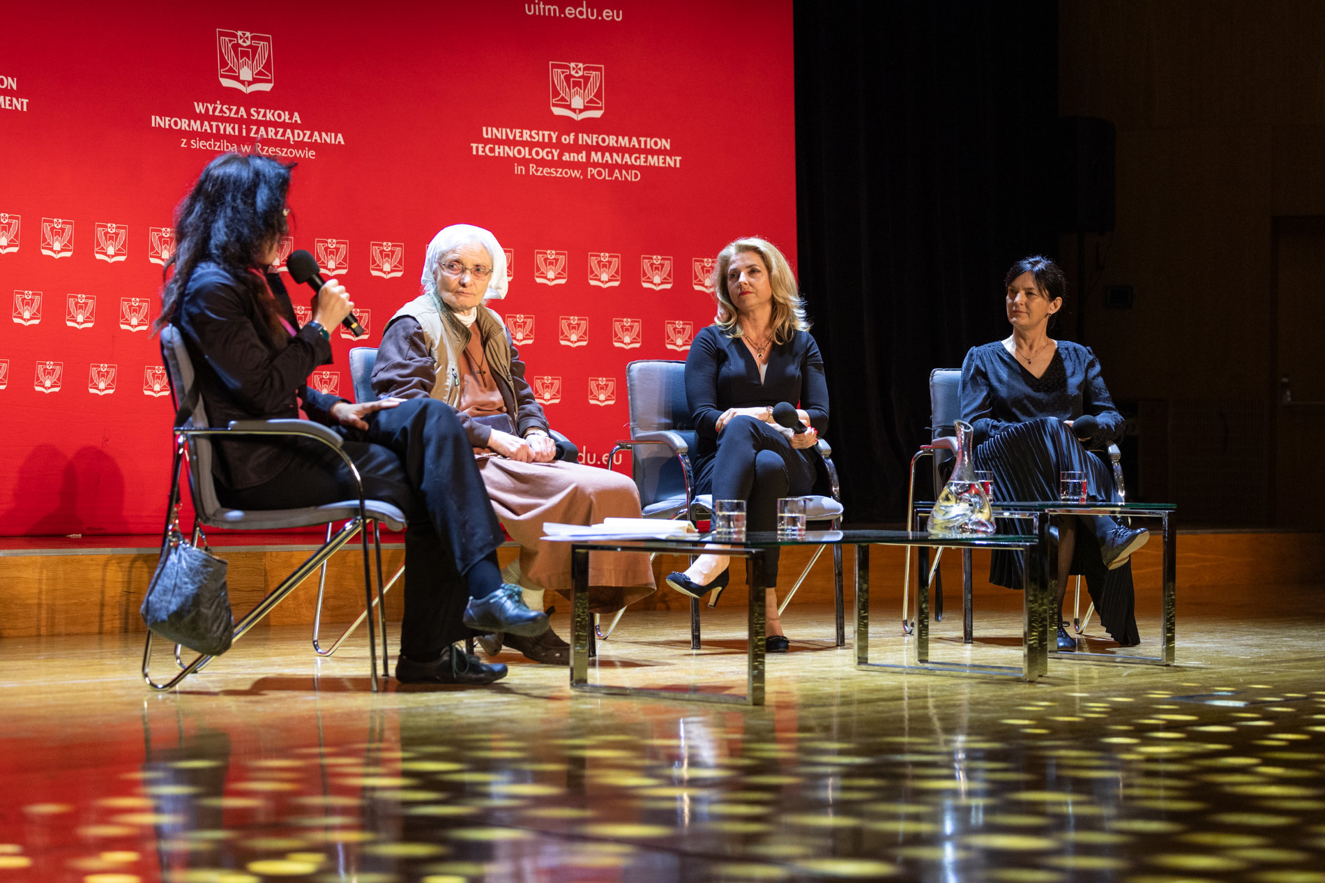 Scena - na krzesłach siedzą cztery kobiety. W tle rozstawiony jest duży, czerwony baner Wyższej Szkoły Informatyki i Zarządzania z siedzibą w Rzeszowie