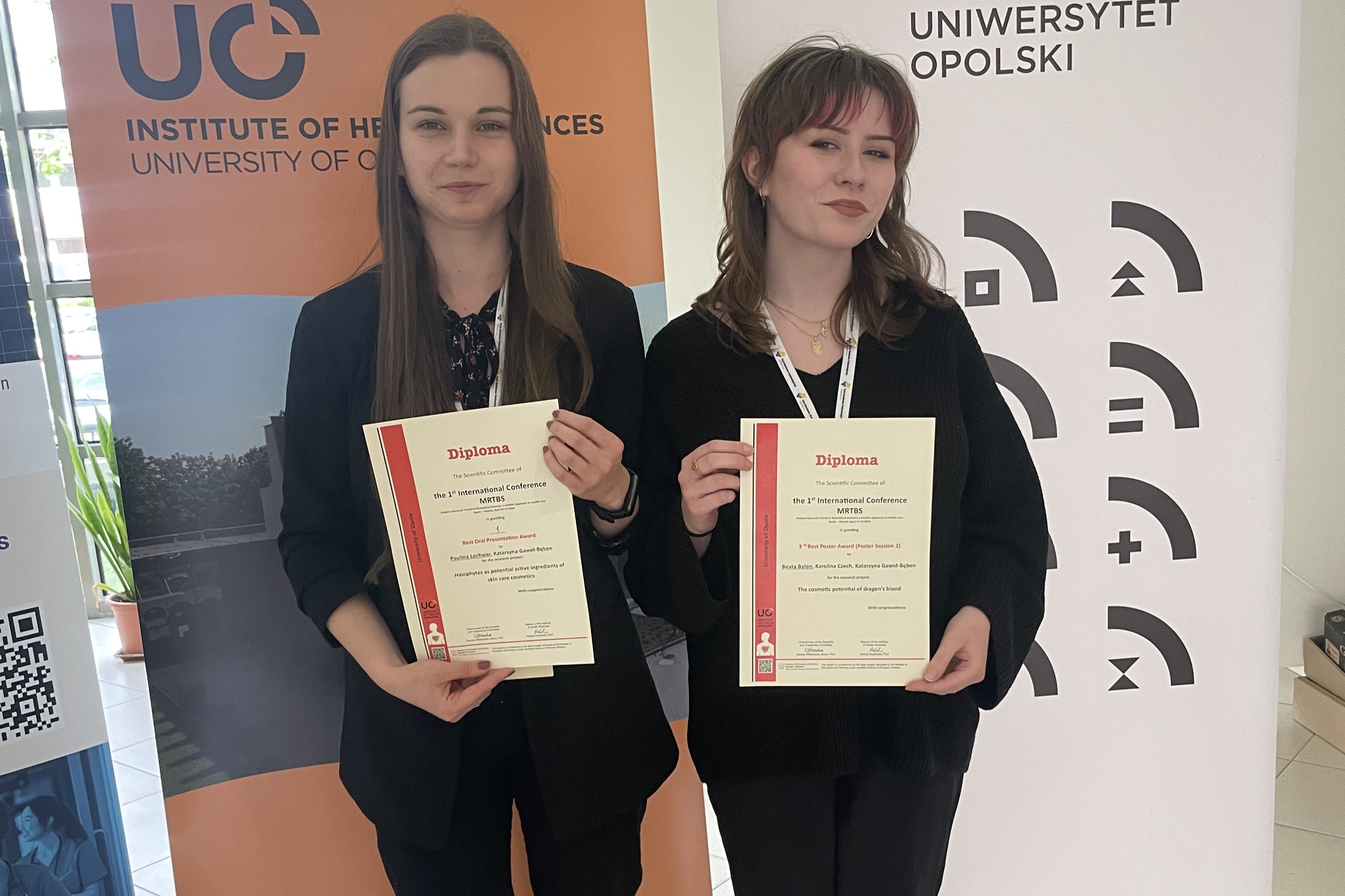 Dwie młode kobiety pozujące do zdjęcia na tle banerów Uniwersytetu Opolskiego. Obie trzymają dyplomy