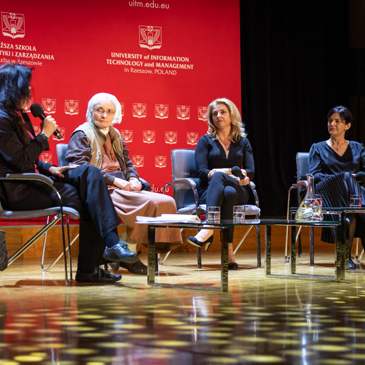 Scena - na krzesłach siedzą cztery kobiety. W tle rozstawiony jest duży, czerwony baner Wyższej Szkoły Informatyki i Zarządzania z siedzibą w Rzeszowie